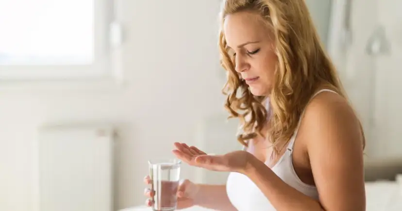 Kobieta z blond włosami bierze tabletkę z dłoni, trzymając szklankę wody.