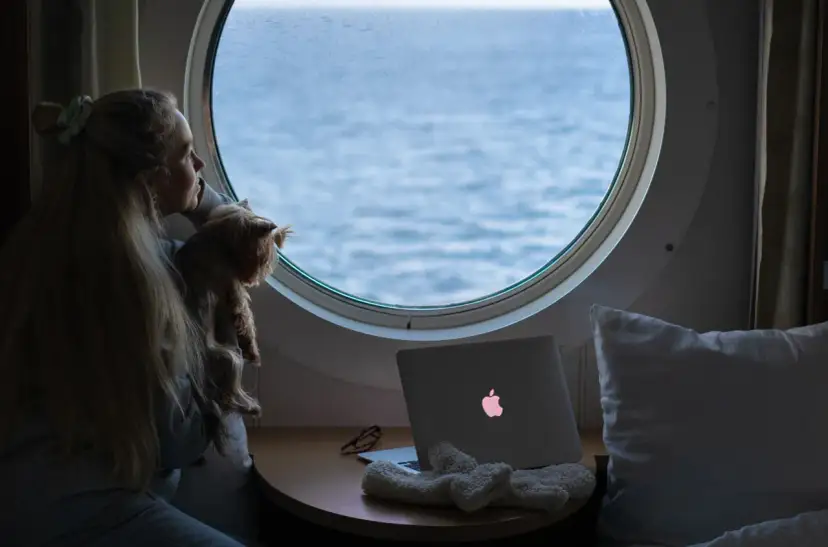 Dziewczyna z psem patrzy przez okrągłe okno na morze. Obok leży laptop z podświetlonym logo, jak prom nova star.