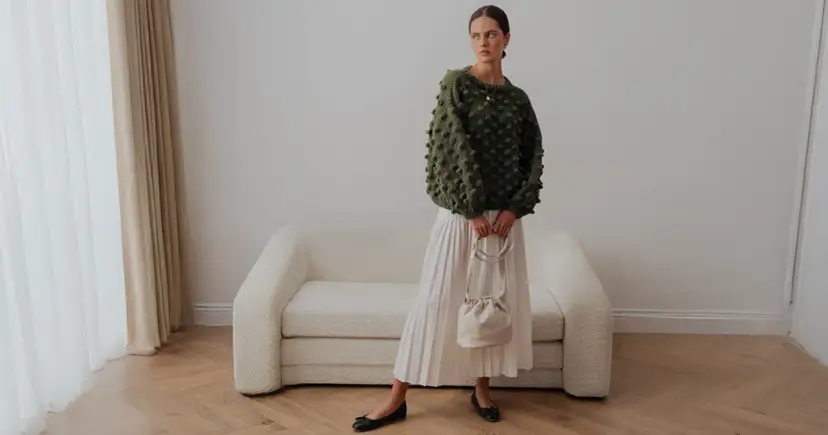 Zielony sweter z wypukłymi kulkami to idealna odpowiedź na pytanie, jaki sweter do letniej sukienki. Stylizacja z białą spódnicą i torebką.
