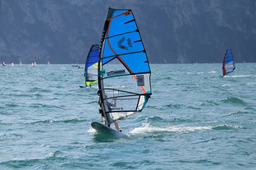 Osoba na desce windsurfingowej z niebieskim żaglem na wzburzonym morzu.