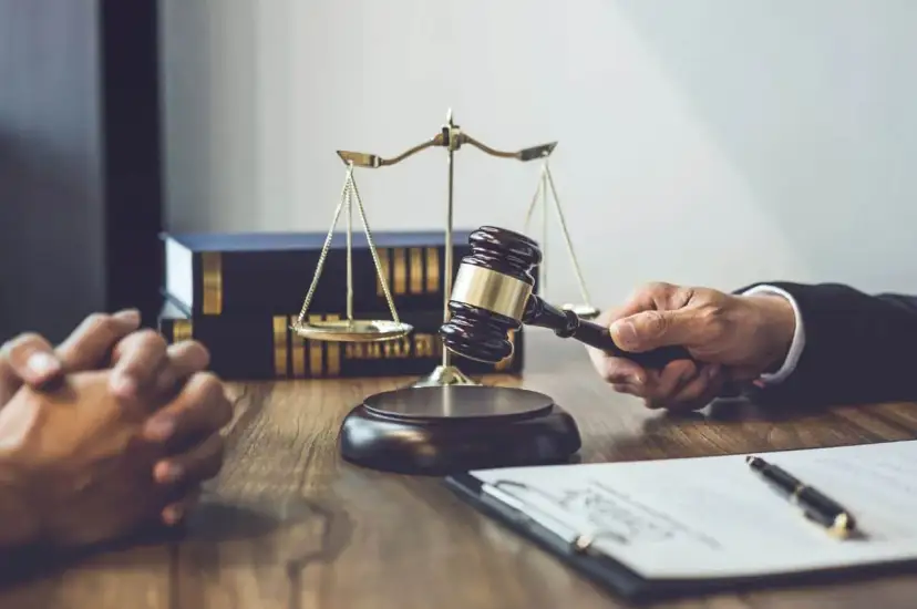 Dłoń trzymająca młotek sędziowski, waga sprawiedliwości i stos książek prawniczych na drewnianym stole.