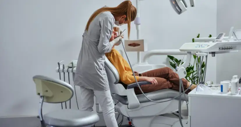 Dentystka w masce i rękawiczkach wykonuje zabieg pacjentowi siedzącemu na fotelu dentystycznym.