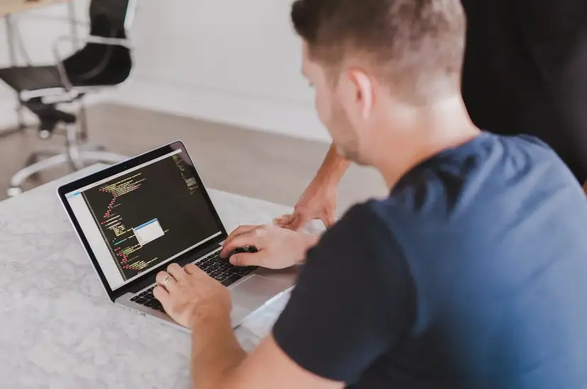 Mężczyzna pisze kod na laptopie, drugi mężczyzna patrzy.