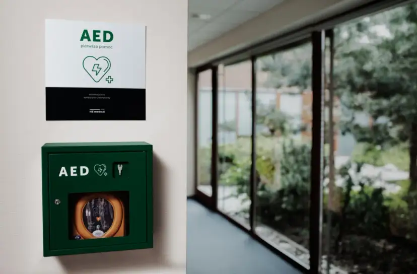 Zielona szafka z defibrylatorem AED na ścianie, obok tabliczka informacyjna.