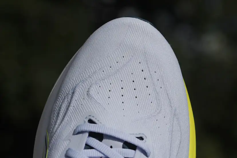 Zbliżenie na jasnoszarą, teksturowaną cholewkę buta sportowego z widocznymi perforacjami i sznurówkami.