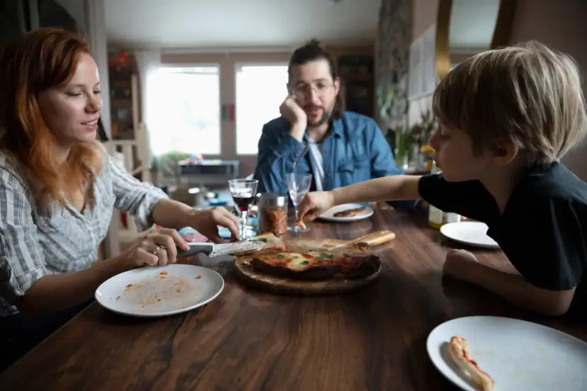 Rodzina przy stole, pizza, w tle mężczyzna zamyślony. Czasem nawet przy wspólnym posiłku pojawiają się kłótnie o dzieci z poprzedniego związku.