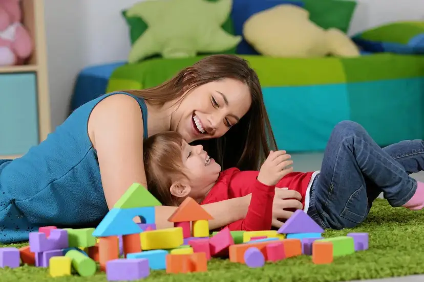 Uśmiechnięta opiekunka bawi się klockami z rocznym dzieckiem. Zastanawiasz się, ile kosztuje niania do rocznego dziecka?