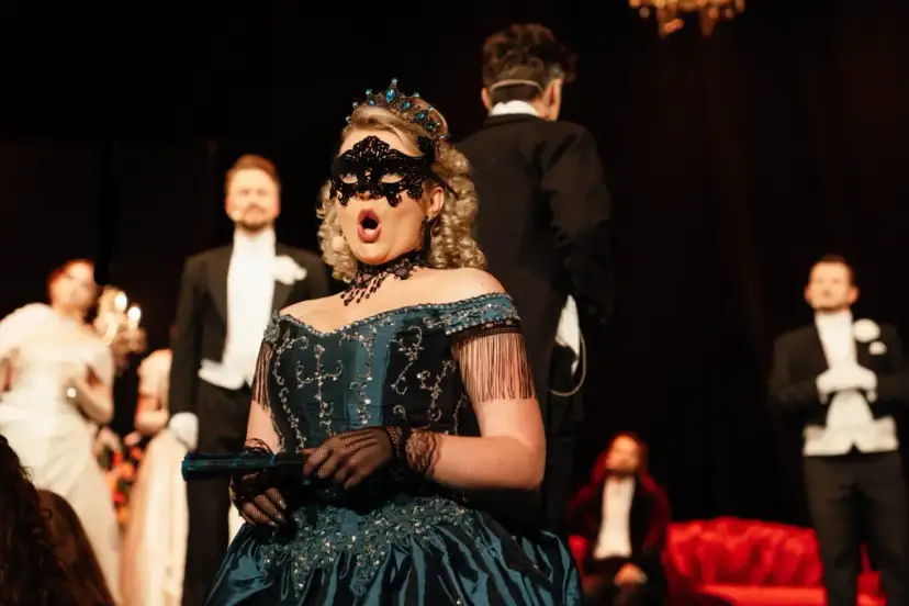 Kobieta w maskę i tiarze śpiewa na scenie. W tle mężczyźni w smokingach. Spektakle w Teatrze Gdynia.