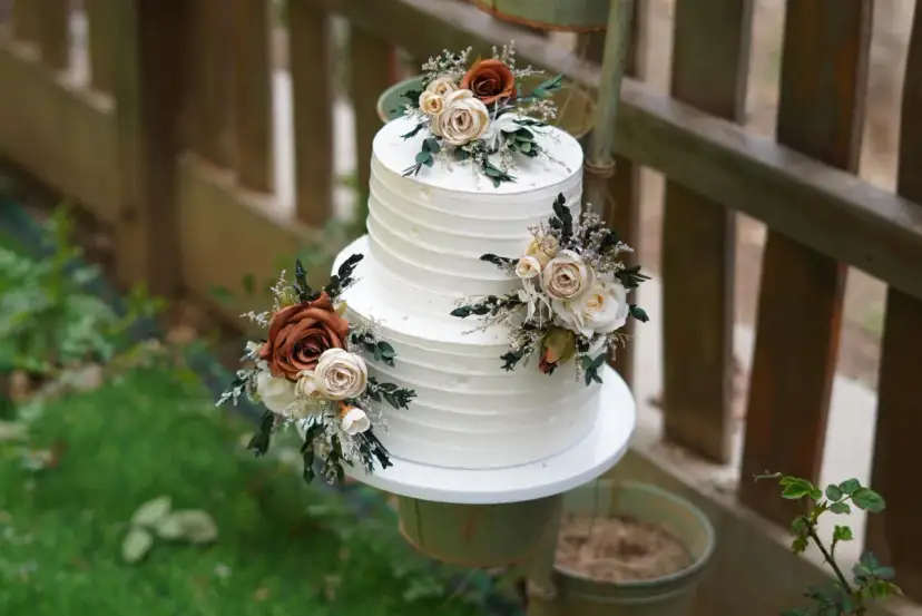 Elegancki tort weselny z białym kremem, ozdobiony bukietami kwiatów w odcieniach brązu i kremu. Wygląda jak tort z margaretkami, idealny na rustykalne przyjęcie.