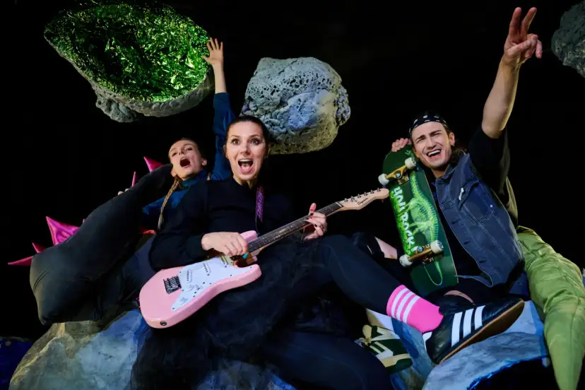 Energiczna ekipa w kosmicznej scenografii, grająca polski folk rock. Kobieta z różową gitarą, mężczyzna ze skateboardem, wesołe miny i podniesione ręce.