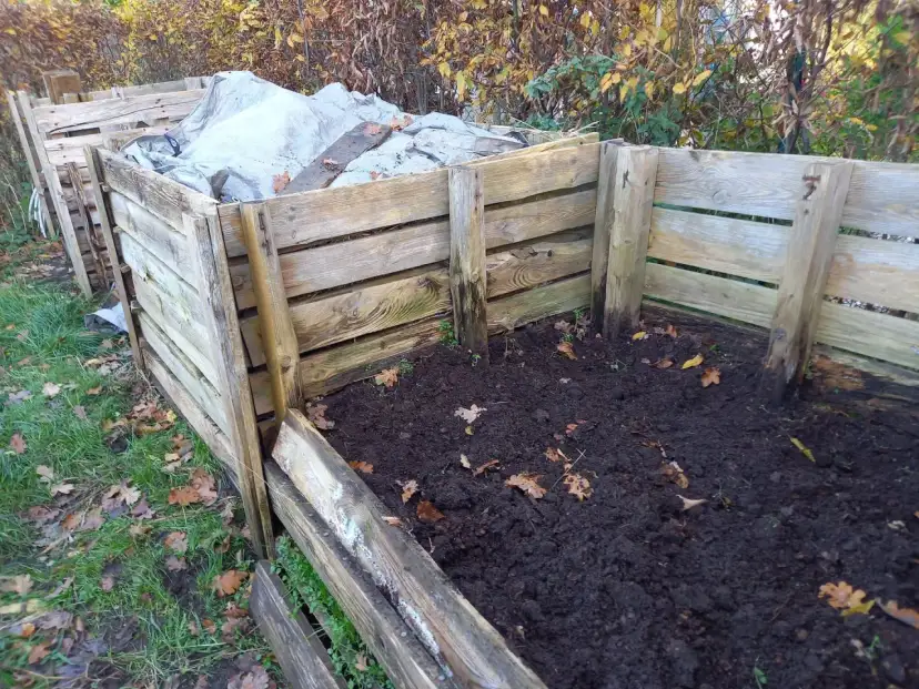 Drewniany kompostownik w ziemi, częściowo przykryty plandeką. Widać jesienne liście i trawę.