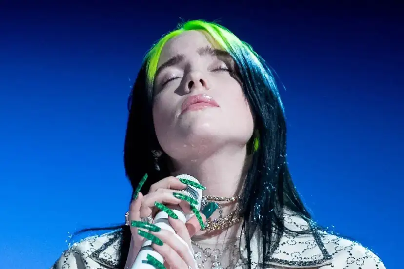 Piosenkarka Billie Eilish z zamkniętymi oczami śpiewa do mikrofonu, jej zielone pasemka włosów lśnią.
