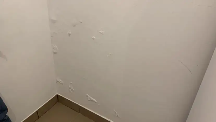 Widoczne pęcherze i łuszcząca się farba na białej, mokrej ścianie w rogu pokoju, wskazujące na problem z wilgocią.