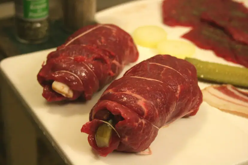 Dwa surowe bitki wołowe zawijane z ogórkiem i boczkiem, gotowe do smażenia.