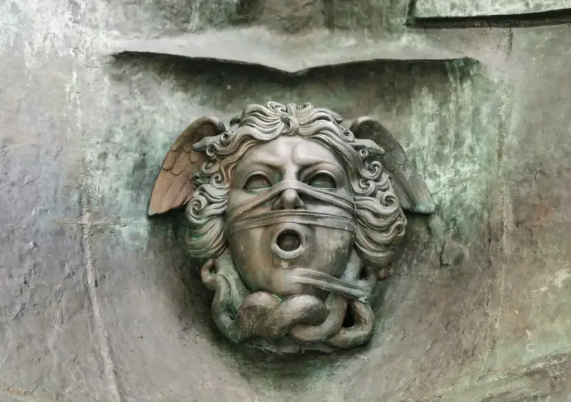 Brązowa rzeźba głowy z opaską na oczach i ustach, ze skrzydłami i wężami.