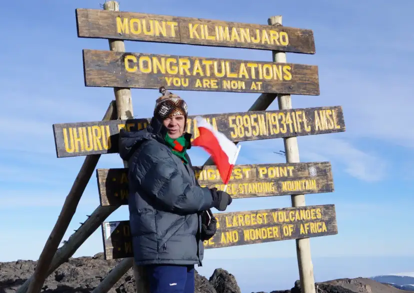 Zdobywca na szczycie **góry Kilimandżaro** z polską flagą. Na tablicy widnieje napis "Congratulations, you are now at Uhuru Peak 5895m".