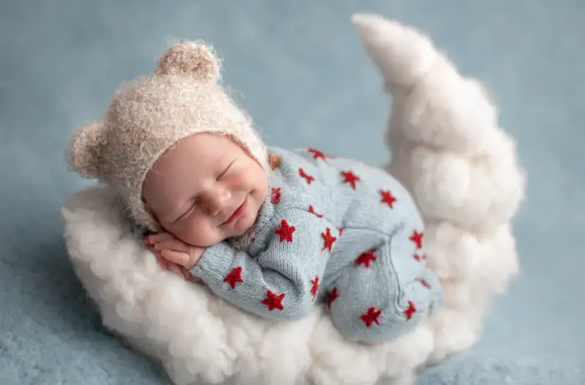 Uśmiechnięte niemowlę w czapce z uszkami śpi na chmurce w kształcie księżyca.