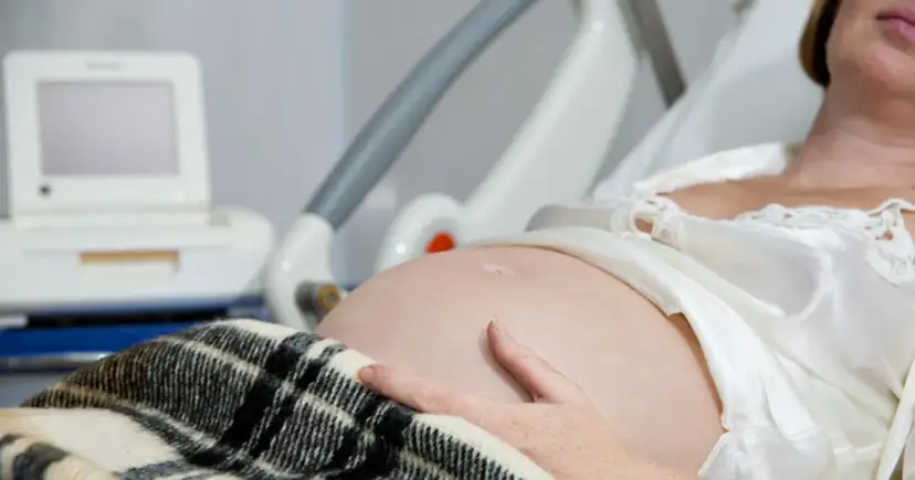 Brzuch kobiety w ciąży, ręka na brzuchu, monitor KTG i łóżko szpitalne.