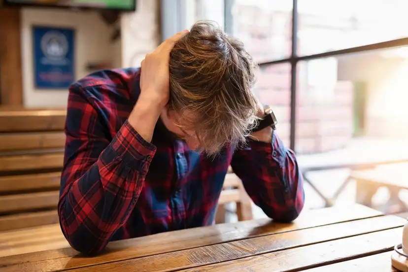 Mężczyzna w kratę z rękami na głowie siedzi przy drewnianym stole.