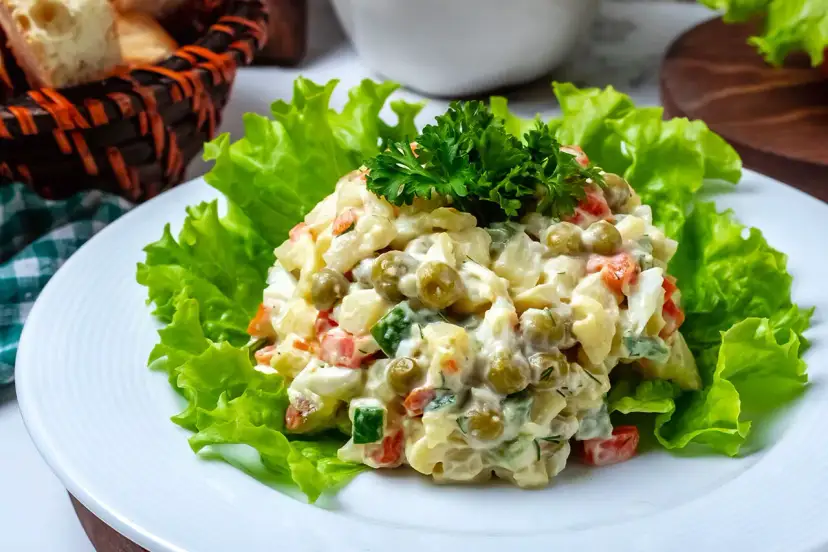 Sałatka jarzynowa z groszkiem, marchewką i ziemniakami, udekorowana natką pietruszki, na liściach sałaty.