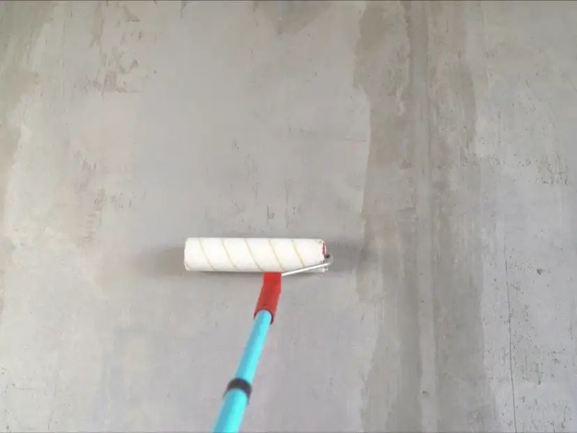 Gruntowanie ścian i sufitów przed tynkowaniem. Wałek malarski z niebieskim trzonkiem nanosi preparat na szarą, betonową powierzchnię.