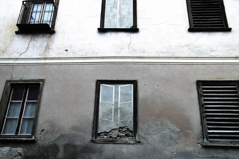 Stara elewacja z widocznymi pęknięciami i zaciekami. Okna z żaluzjami i jedno wybite.
