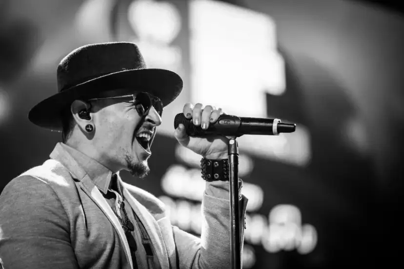Wokalista Linkin Park, Chester Bennington, w kapeluszu i okularach przeciwsłonecznych, śpiewa do mikrofonu z pasją.