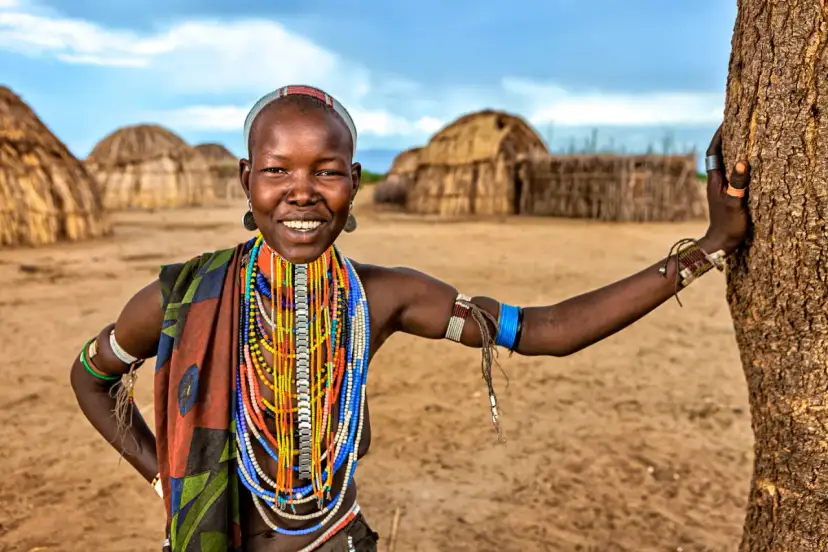 Uśmiechnięta kobieta z Etiopii w tradycyjnych ozdobach, w tle chaty. Niezapomniana etiopia wycieczka.
