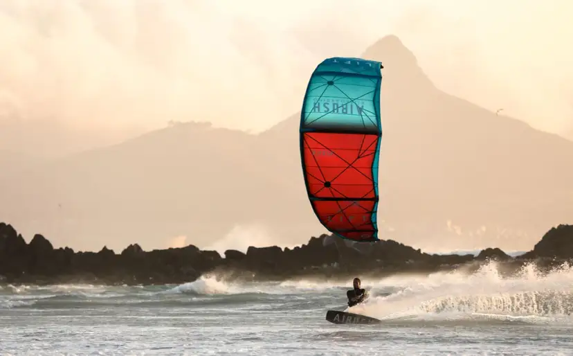 Kitesurfer na desce w akcji, z kolorowym latawcem na tle gór i morza.