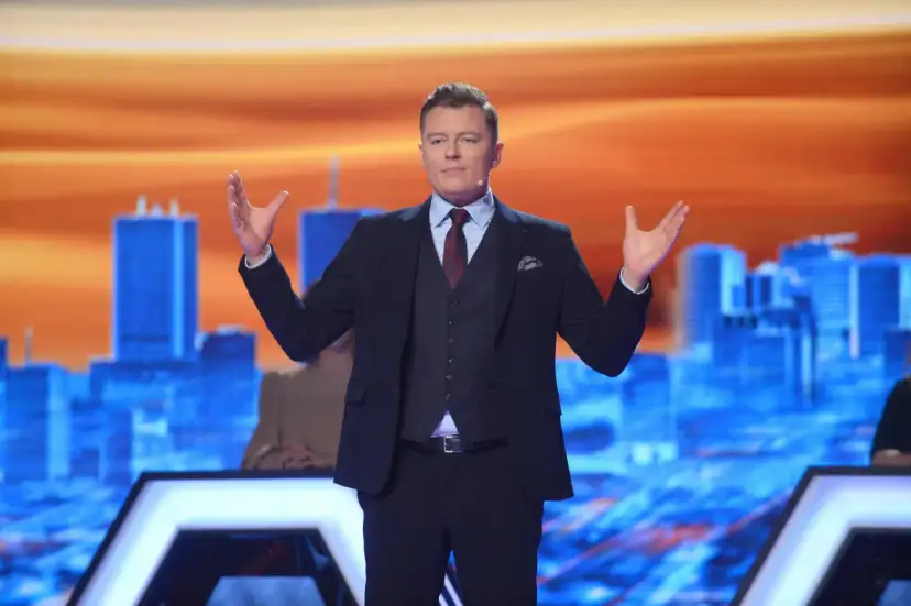 Rafał Brzozowski w garniturze, z rękami uniesionymi na boki, prowadzi program "Jaka to melodia?".