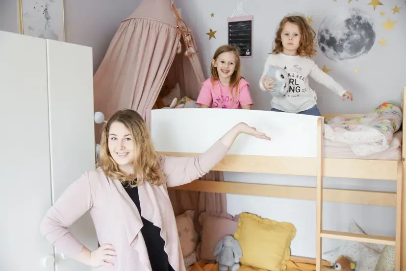 Uroczy mały pokój dla rodzeństwa w bloku. Dwie dziewczynki bawią się na łóżku piętrowym, a mama prezentuje przestrzeń.