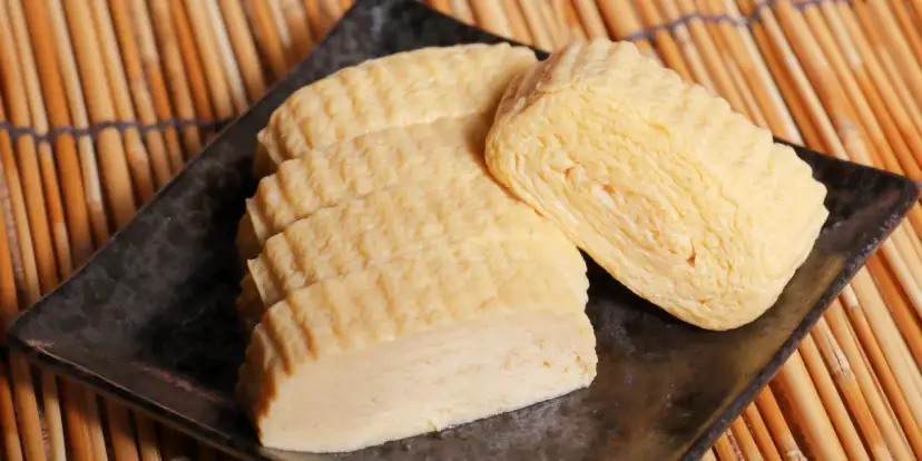 Trzy porcje nigiri tamago, delikatnie zwiniętego omletu jajecznego, ułożone na czarnym talerzu na tle bambusowej maty.