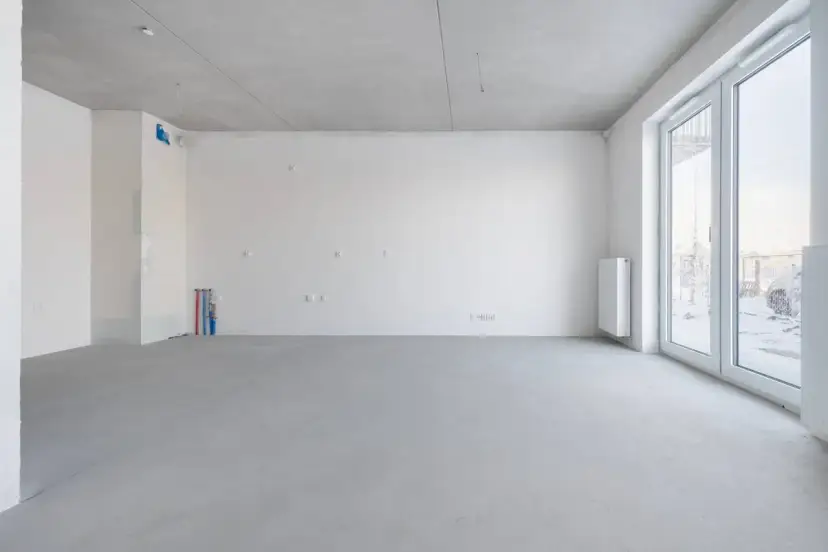Pusta przestrzeń w stanie deweloperskim: betonowa posadzka, białe ściany, instalacje wodne i elektryczne, grzejnik, duże okna.