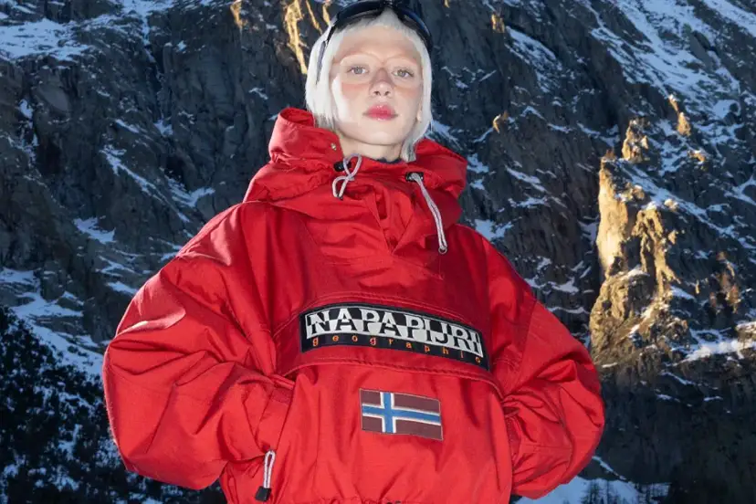 Modelka w czerwonej kurtce z logo Napapijri Geographic i flagą Norwegii na tle ośnieżonych gór. Napapijri co to za firma? To marka odzieży outdoorowej.