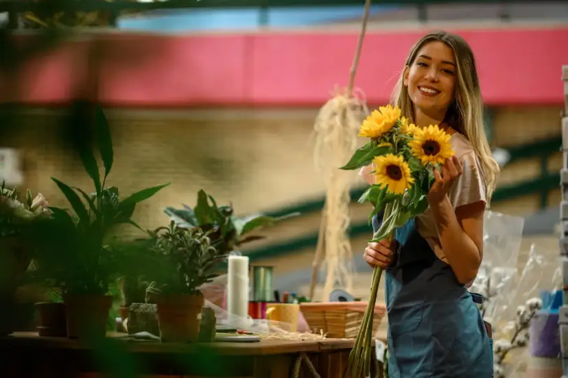 Uśmiechnięta młoda kobieta w fartuchu trzyma bukiet słoneczników. To idealny obrazek dla kogoś, kto zastanawia się, jak zostać florystą.