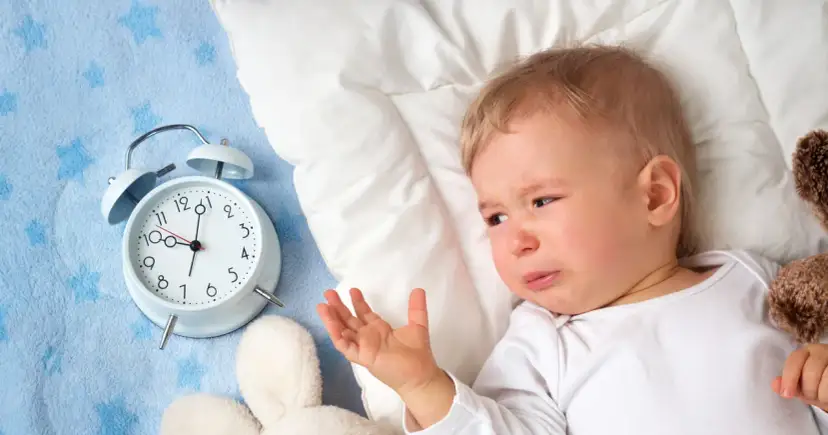 Płacz dziecka obok budzika sugeruje, dlaczego dziecko nie chce spać. Maluch leży na niebieskim kocyku z gwiazdkami, obok pluszowych zabawek.