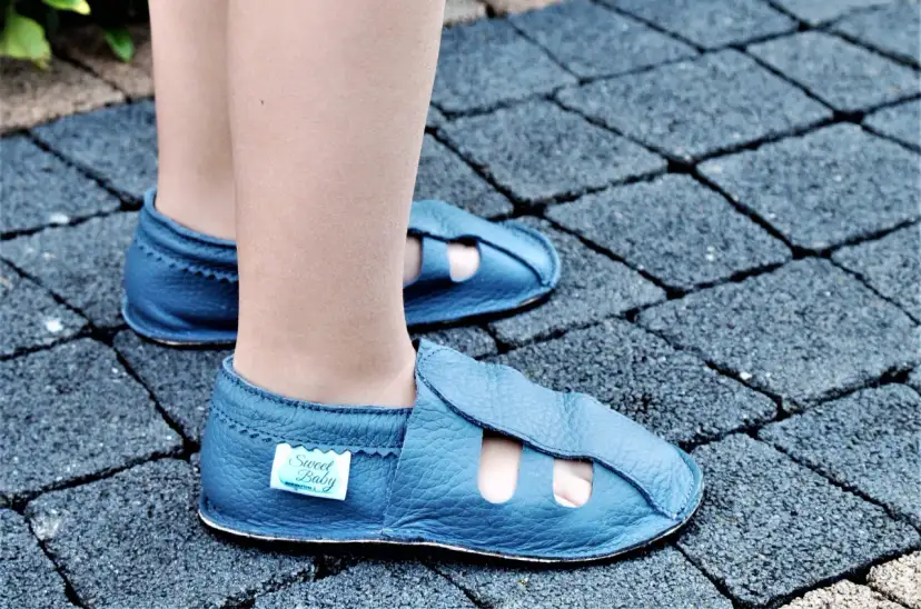 Niebieskie skórzane kapcie dziecięce z otworami na palce na tle kostki brukowej.