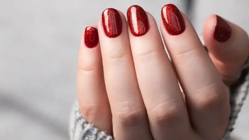 Glitzernde rote Gel-Nagel-Designs auf einer Hand, die aus einem grauen Pullover ragt.