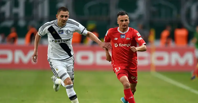 Dwóch piłkarzy w akcji. Jeden w białej koszulce Legii, drugi w czerwonej. Ich gra to dowód na najwyższe zarobki w ekstraklasie.