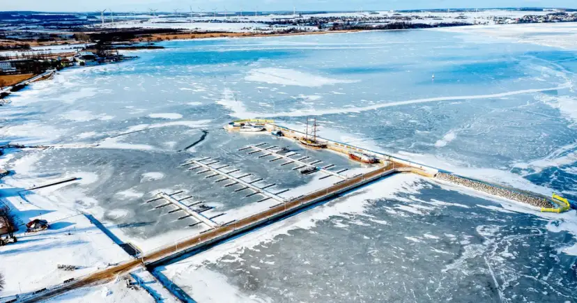 Zamarznięte morze bałtyckie zimą. Widok na zamarzniętą zatokę z przystanią, statkami i wiatrakami na horyzoncie.