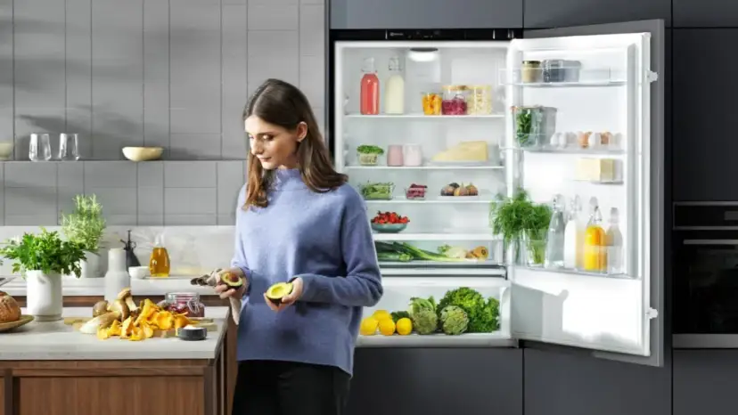 Kobieta kroi awokado obok otwartej lodówki pełnej świeżych produktów.