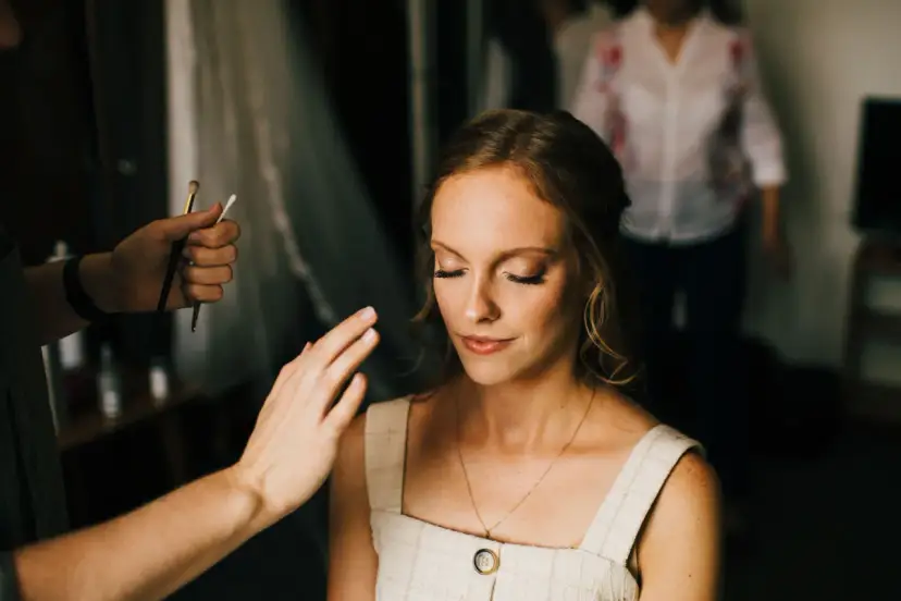 Kobieta z zamkniętymi oczami, której makijaż jest poprawiany przez wizażystkę.
