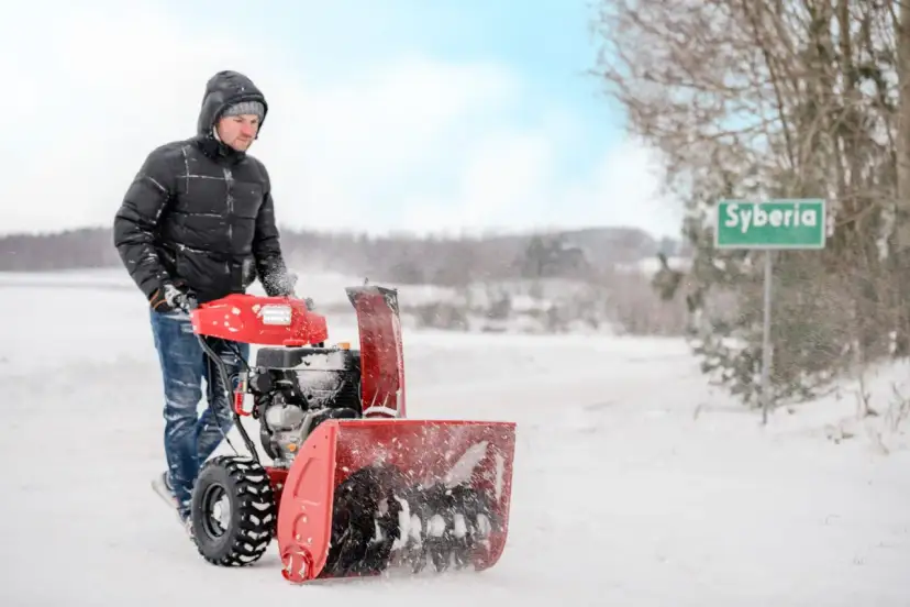 Mężczyzna w zimowej kurtce używa odśnieżarki do śniegu, by oczyścić drogę. W tle zielony znak z napisem "Syberia".