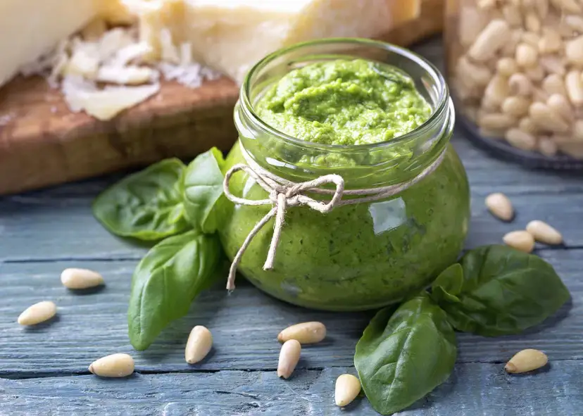 Słoik domowego pesto z bazylii, otoczony świeżymi liśćmi bazylii i orzeszkami pinii. Obok kawałki parmezanu. Idealne do makaronu.