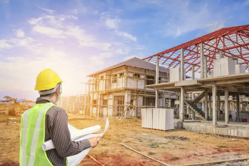 Inżynier w żółtym kasku i kamizelce odblaskowej analizuje plany budowy domu. Czy to pozwolenie na budowę czy zgłoszenie?