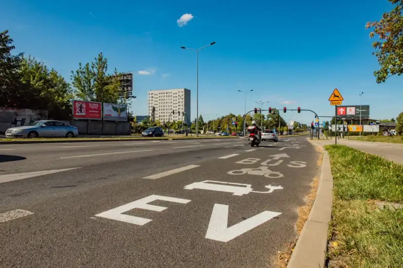 Na drodze oznaczenie "EV BUS" sugeruje, czy samochody elektryczne mogą jeździć buspasem. W tle budynek i ruch uliczny.