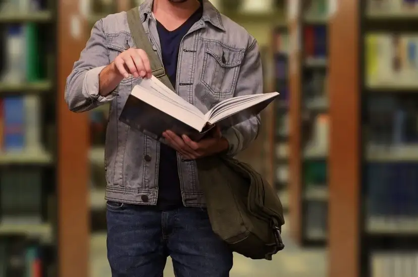 Student w dżinsowej kurtce przegląda książkę w bibliotece. W tle widać regały z książkami.