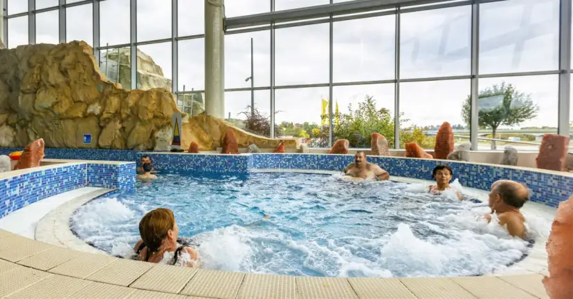Ludzie relaksują się w basenie termalnym z hydromasażem, otoczonym skalnymi formacjami i dużymi oknami.