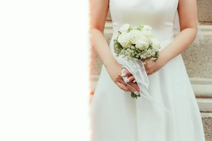 Panna młoda trzyma bukiet serc z białych róż i gipsówki, ozdobiony białą wstążką.