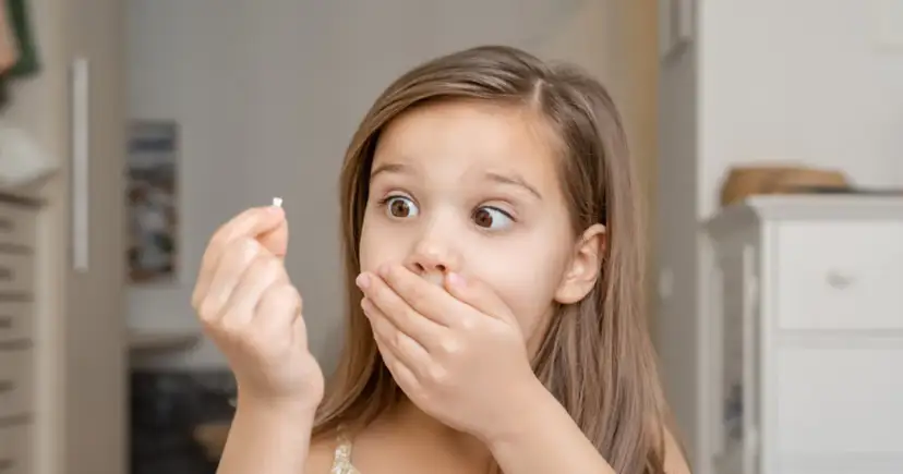 Zaskoczona dziewczynka trzyma w dłoni mały mleczak, zastanawiając się, kiedy wypadają mleczaki.